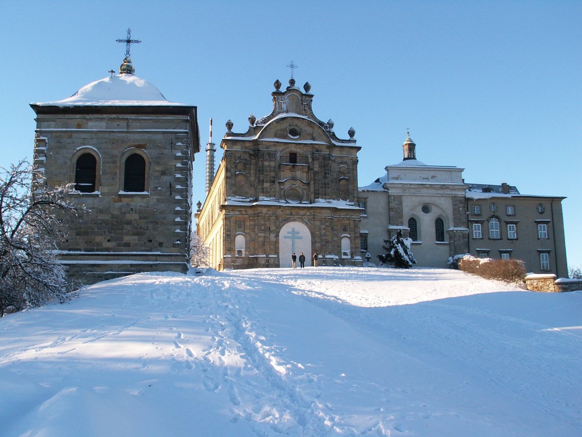 Exterior view of the Holy Cross Benedictine monastery complex in Poland under snow, with bell tower and chapel facade