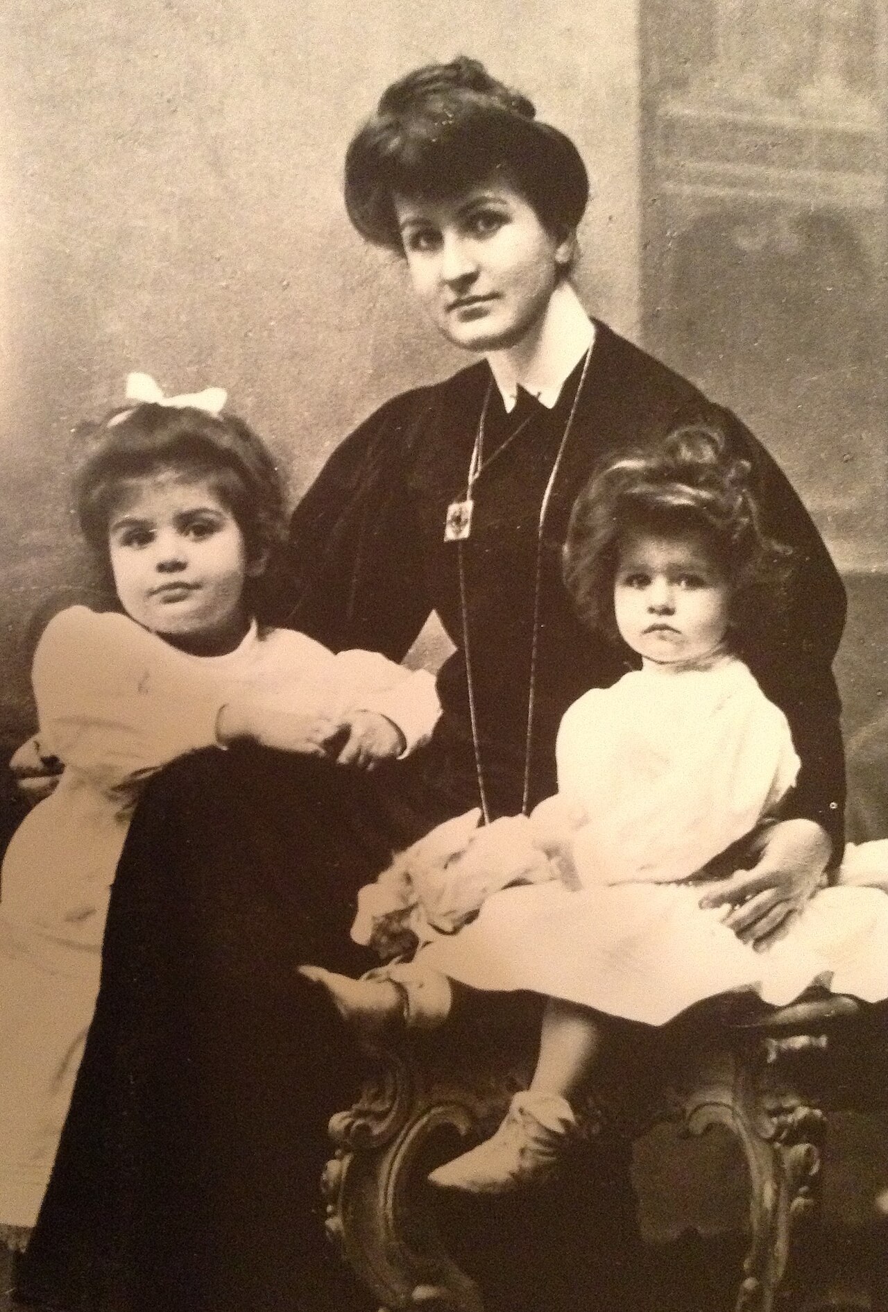 Sepia photograph of Alma Mahler in a dark dress seated with her two young daughters Maria and Anna