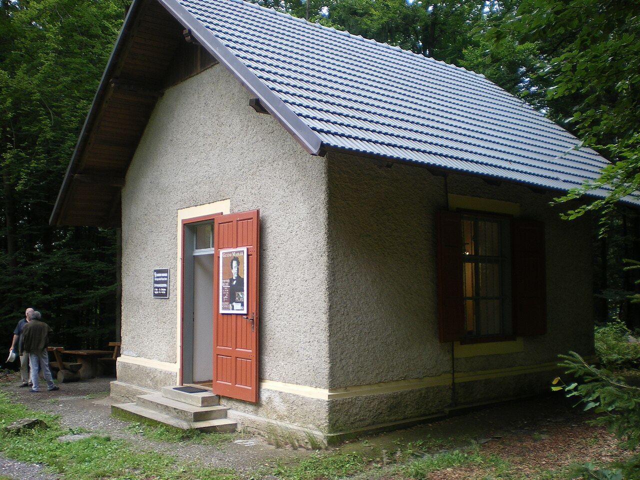 Small stone composing hut with a tiled roof standing among trees, with a poster of Gustav Mahler fixed to its door
