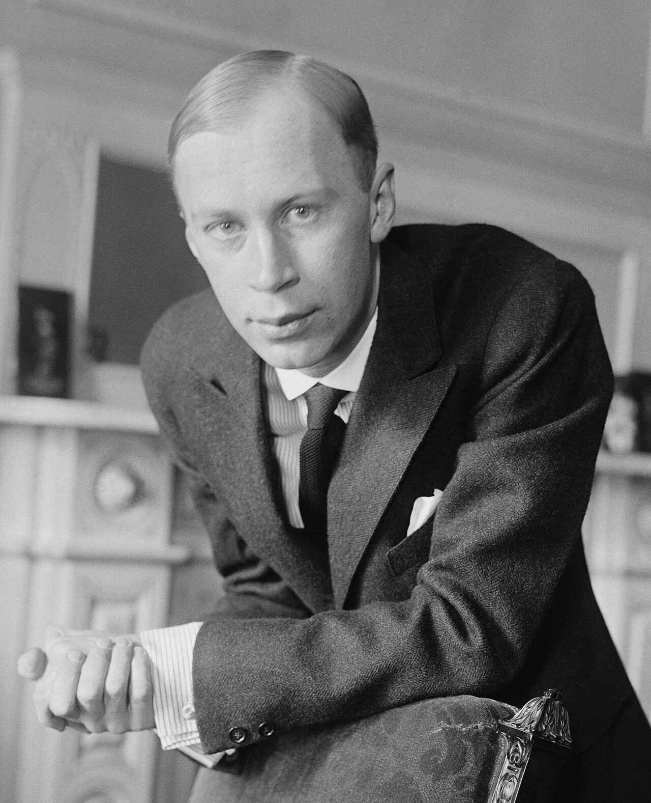 Black-and-white portrait photograph of the young composer Sergei Prokofiev in a suit, seated beside a chair, around 1918