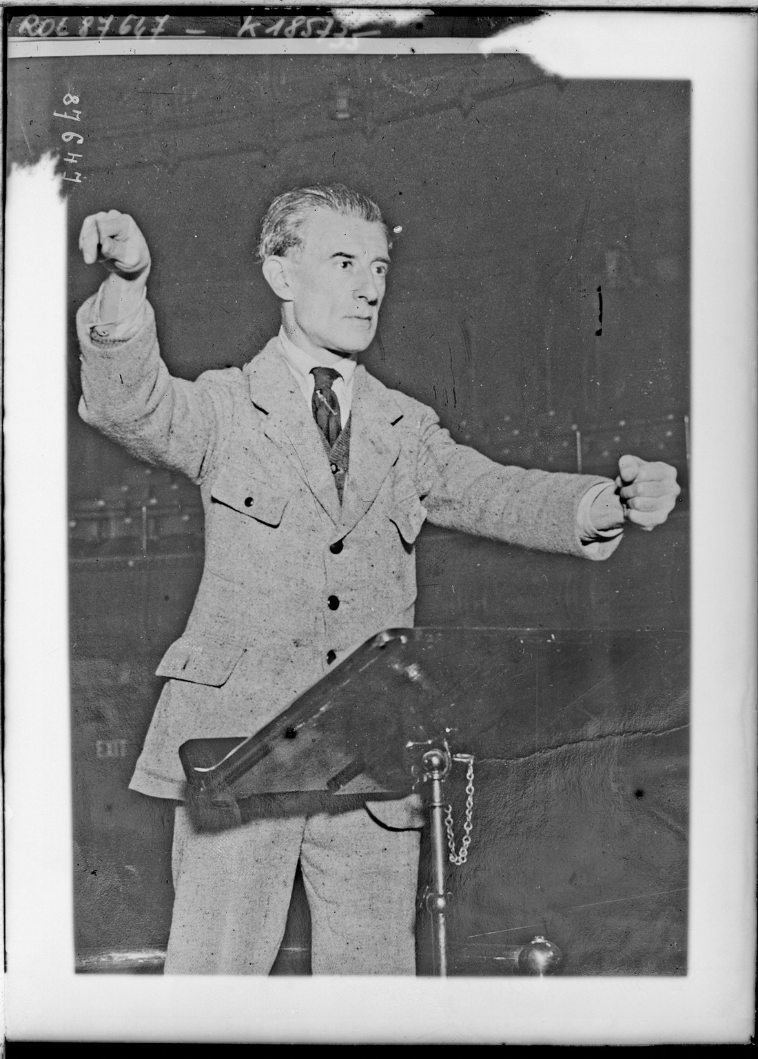 Black-and-white press photograph of Maurice Ravel conducting an orchestra with both arms raised